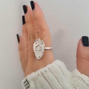 Könnte beinhalten: Ein silberner Ring mit einem skulpturalen Gesichtsmotiv und detaillierten Haarverzierungen am oberen Rand. Das Gesicht weist ausgeprägte Augenbrauen, eine Nase und einen kleinen Mund auf, ergänzt durch eine glatte Ringschiene aus poliertem Metall.