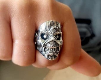May include: A silver-toned metal ring featuring a detailed, textured skull design with deep-set eye sockets, a prominent brow ridge, and bared teeth. The surface of the ring has a weathered, metallic finish with dark recessed areas to highlight the features.