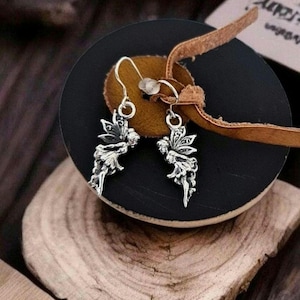 Puede incluir: Un par de pendientes de tono plateado con un diseño detallado de hada con alas extendidas. Los pendientes cuelgan de finos ganchos de metal y muestran texturas intrincadas en las figuras de hadas, exhibiendo un acabado metálico.