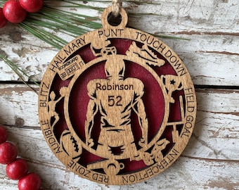 May include: A wooden Christmas ornament featuring a football player silhouette with the name "Robinson" and the number "52" on the back. The ornament is decorated with various football-related terms, including "Touchdown", "Punt", "Hail Mary", "Red Zone", "Interception", "Field Goal", "Blitz", and "Audible".