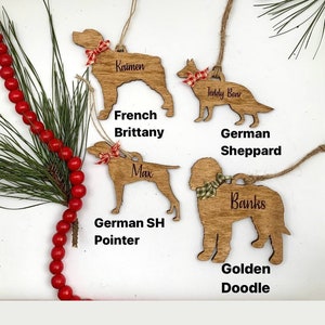 May include: Five wooden dog breed ornaments with personalized names. The ornaments are in the shapes of a French Brittany, German Shorthaired Pointer, Golden Doodle, German Shepherd, and Teddy Bear. Each ornament has a red and white or green and white bow.