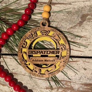 May include: A wooden ornament with a yellow center and the word "DISPATCHER" in a banner. The ornament is decorated with the words "Dedicated, Dependable, Helpful, Efficient, Reliable, Hard Working, Multitasker". The name "Addison Metcalf" is written on a small banner below the word "DISPATCHER".