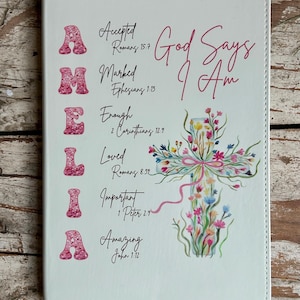 May include: White journal with the name "AMELIA" in pink glitter letters. The journal features the text "God Says I Am" and inspirational phrases with Bible verses, along with a floral cross design.