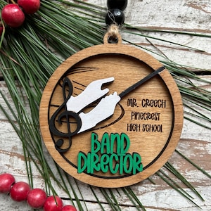 May include: A wooden Christmas ornament with a green and black design. It features a musical note, a conductor's baton, and the text "Band Director". The ornament also includes the text "Mr. Creech Pinecrest High School".
