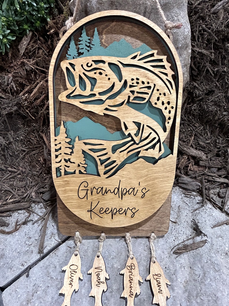 May include: Wooden wall hanging with a cutout of a bass fish jumping out of the water. The hanging has the text "Grandpa's Keepers" and five fish-shaped tags hanging below with the names Oliver, Noah, Brianna, Liam, and Lucy.