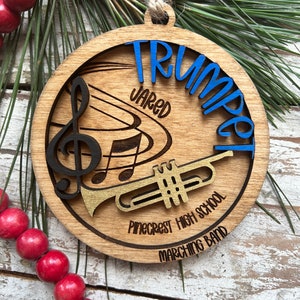 May include: A wooden Christmas ornament with a trumpet design. The ornament is painted in gold, black, and blue. The text "Trumpet" is written in blue, and the text "Jared Pinecrest High School Marching Band" is written in black.
