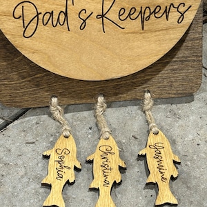 May include: Wooden sign with the text "Dad's Keepers" and three wooden fish-shaped tags hanging from it. The tags are labeled "Sophia", "Christina", and "Jasmine".