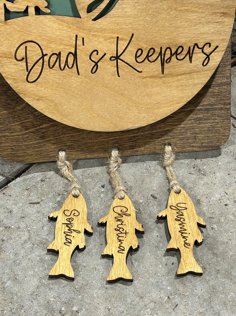 May include: Wooden sign with the text "Dad's Keepers" and three wooden fish-shaped tags hanging from it. The tags are labeled "Sophia", "Christina", and "Jasmine".