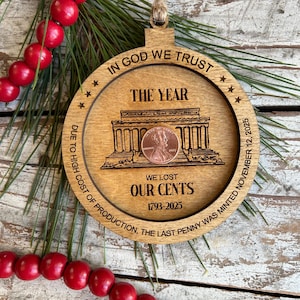 May include: Wooden Christmas ornament with a penny inside, featuring the text "IN GOD WE TRUST" and "WE LOST OUR CENTS 1793-2025". The ornament has a circular design with a building illustration and the words "DUE TO HIGH COST OF PRODUCTION, THE LAST PENNY WAS MINTED NOVEMBER 12, 2025".