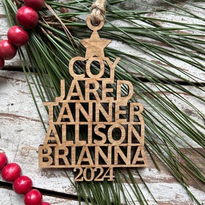 May include: A wooden Christmas tree ornament with the names Coy, Jared, Tanner, Allison, and Brianna, and the year 2024 cut out. The ornament is suspended by a twine loop and rests on a bed of pine needles and red berries.