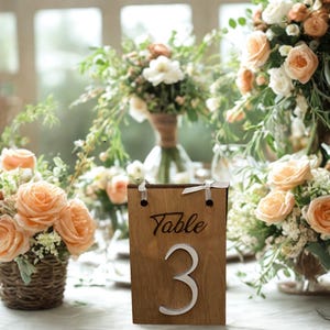 May include: A wooden table number 3 with the word "Table" engraved on it, displayed on a table set for a wedding. Peach roses and greenery are in the background, creating a soft, romantic atmosphere.