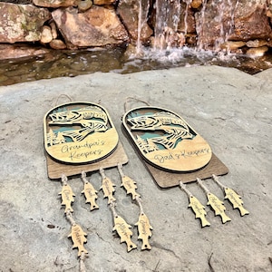 May include: Two wooden fish-shaped wall hangings with the text "Grandpa's Keepers" and "Dad's Keepers" on them. Each hanging has a string of smaller fish-shaped tags with names written on them.
