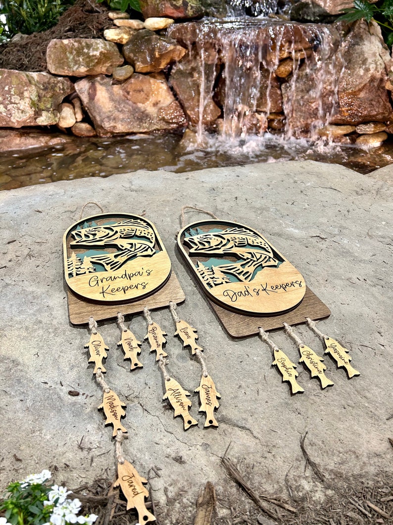 May include: Two wooden fish-shaped wall hangings with the text "Grandpa's Keepers" and "Dad's Keepers" on them. Each hanging has a string of smaller fish-shaped tags with names written on them.