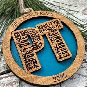 May include: Wooden Christmas ornament with a teal blue center and a brown wooden frame. The ornament features the letters "PT" with words related to physical therapy, the name "DR. TANNER REVIS", and the year "2025". A twine loop is attached for hanging.