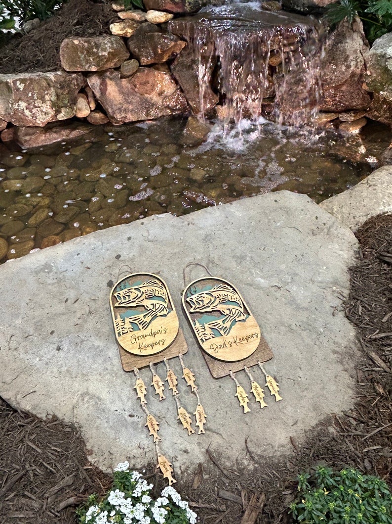 May include: Two wooden fish-shaped wall hangings with the text "Grandpa's Keepers" and "Dad's Keepers" on them. Each hanging has a string of smaller fish-shaped charms hanging from it.