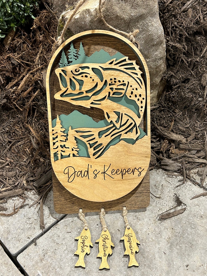May include: Wooden wall hanging with a carved bass fish and the text "Dad's Keepers". Three smaller fish-shaped tags hang below with the names "Sophia", "Christina", and "Yasmine" engraved on them.