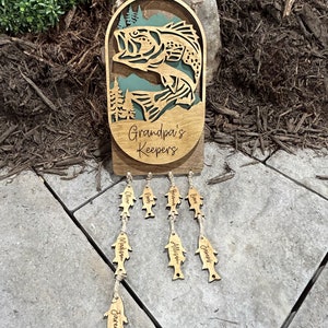 May include: Wooden sign with a bass fish jumping out of the water, surrounded by trees. The sign says "Grandpa's Keepers" and has wooden fish hanging below with names on them: Jared, Madison, Allison, James, Noah, Olivia, and Graham.