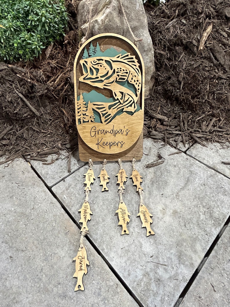 May include: Wooden sign with a bass fish jumping out of the water, surrounded by trees. The sign says "Grandpa's Keepers" and has wooden fish hanging below with names on them: Jared, Madison, Allison, James, Noah, Olivia, and Graham.