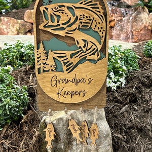 May include: Wooden fish-shaped ornament with the text "Grandpa's Keepers" and smaller fish-shaped ornaments with names including "Oliver", "Noah", "Brianna", "Liam", "Madison", "Alison", and "Emma" hanging from it.
