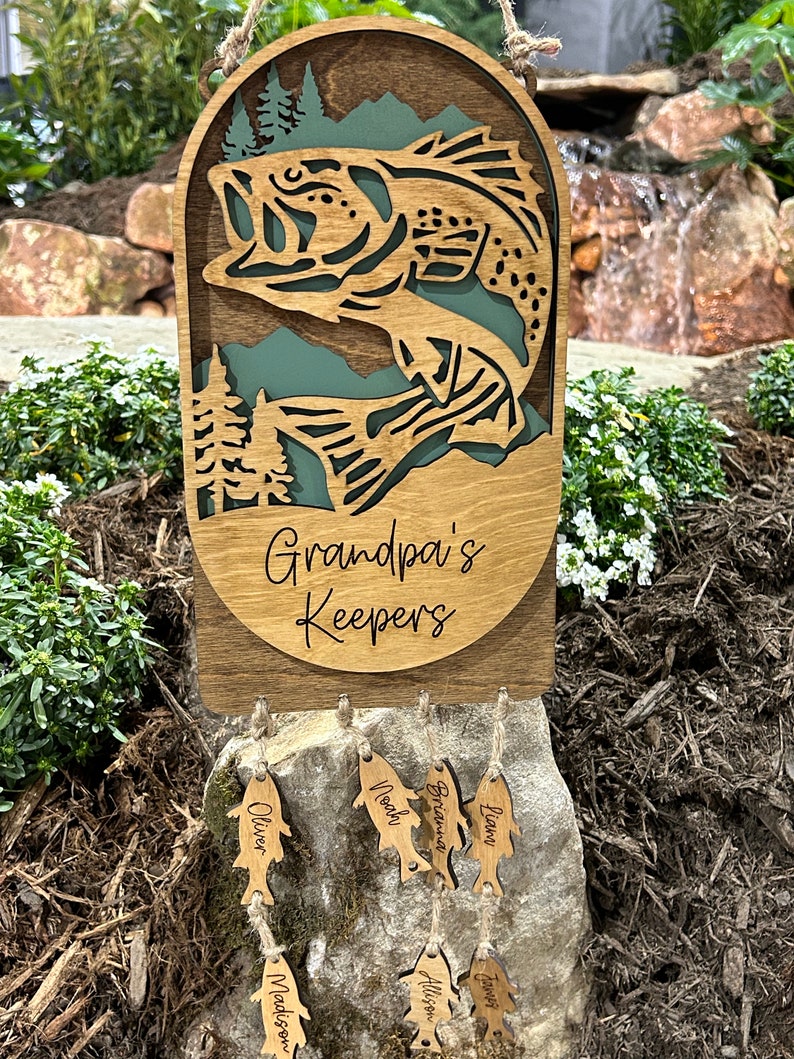May include: Wooden fish-shaped ornament with the text "Grandpa's Keepers" and smaller fish-shaped ornaments with names including "Oliver", "Noah", "Brianna", "Liam", "Madison", "Alison", and "Emma" hanging from it.