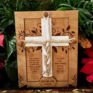 May include: Wooden framed cross with a white cross and rope design. The frame has engraved floral accents and text that reads "They are no longer two, but one flesh..." and "A cord of three strands is not easily broken." Includes names and a date.