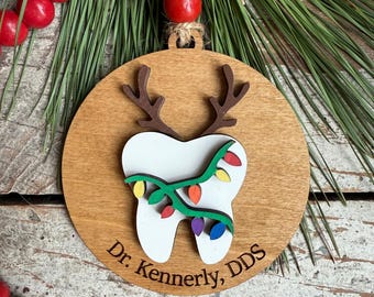 May include: A round wooden ornament featuring a white tooth cutout with brown reindeer antlers and a string of colorful Christmas lights wrapped around it. The text Dr. Kennerly, DDS is engraved at the bottom. It hangs from a jute twine loop.