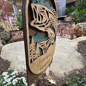 May include: A wooden sign with a cutout of a bass fish jumping out of the water. The sign says "Grandpa's Keepers" and has several smaller wooden tags hanging from it with names written on them.