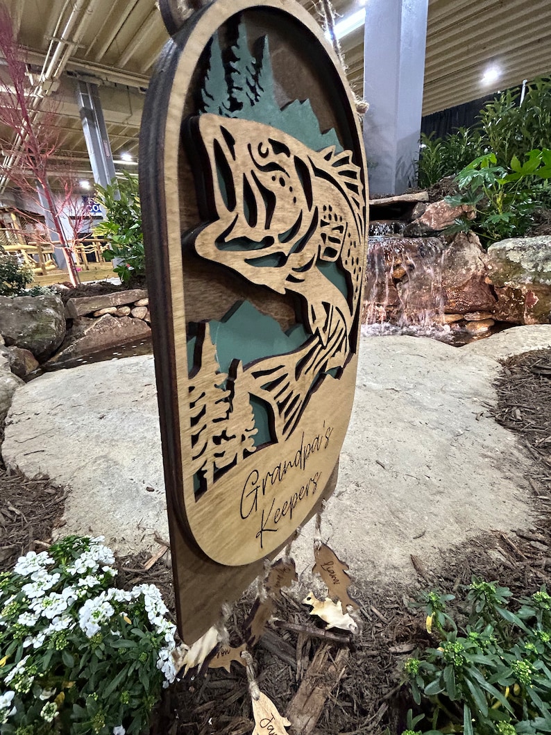 May include: A wooden sign with a cutout of a bass fish jumping out of the water. The sign says "Grandpa's Keepers" and has several smaller wooden tags hanging from it with names written on them.