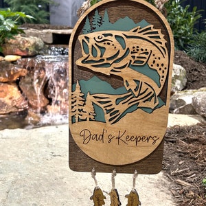 May include: A wooden wall hanging with a bass fish cutout and the text "Dad's Keepers". Three smaller wooden fish hang below with the names "Sophia", "Jackson", and "Benjamin" engraved on them.