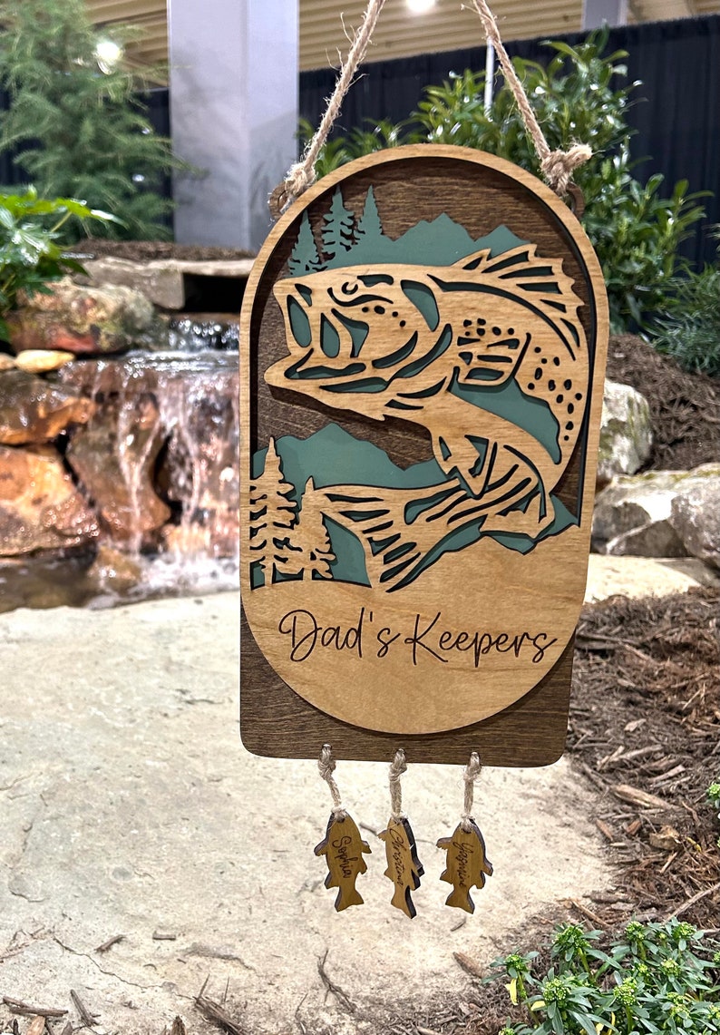 May include: A wooden wall hanging with a bass fish cutout and the text "Dad's Keepers". Three smaller wooden fish hang below with the names "Sophia", "Jackson", and "Benjamin" engraved on them.