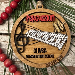 May include: A wooden Christmas ornament with a xylophone, drumsticks, and a treble clef. The ornament is personalized with the name "Oliver" and the text "Newsville High School Marching Band".