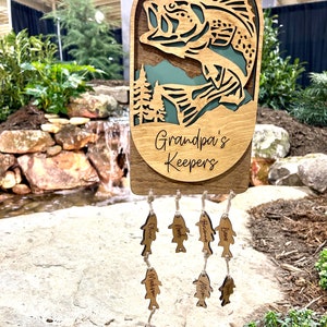 May include: A wooden fish-shaped wind chime with the text "Grandpa's Keepers" on the top. The chime has several smaller fish-shaped charms with names on them, including "Oliver", "Noah", "Brieland", "Liam", "Madisyn", "Allison", "Tyler", and "Ezra".