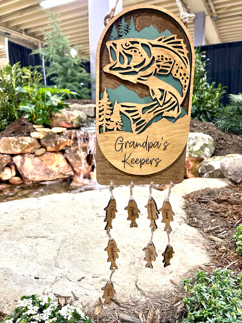 May include: A wooden fish-shaped wind chime with the text "Grandpa's Keepers" on the top. The chime has several smaller fish-shaped charms with names on them, including "Oliver", "Noah", "Brieland", "Liam", "Madisyn", "Allison", "Tyler", and "Ezra".