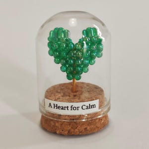 Pode incluir: Uma cúpula de vidro com base de cortiça abriga uma escultura em forma de coração feita de contas verdes. A escultura é montada em um fio dourado. Uma etiqueta na base diz "A Heart for Calm."