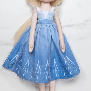 Puede incluir: Muñeca hecha a mano con pelo largo rubio y un vestido azul degradado. El vestido presenta un corpiño blanco, tirantes finos y una falda con un patrón geométrico blanco. Una superposición de tul blanco añade volumen.