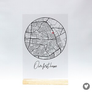 Op de afbeelding: Een zwart-witte kaart van een stad met een rood hart dat een locatie markeert. De kaart is gedrukt op een heldere acrylplaat met een houten standaard. De tekst "Our first home" is onder de kaart gedrukt.