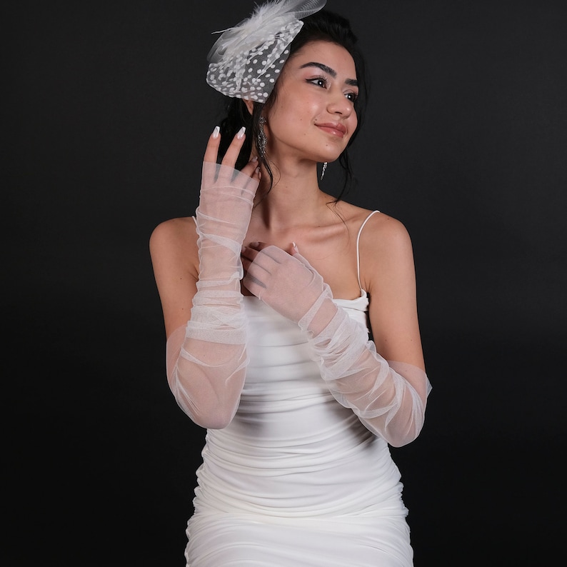 May include: A white dress with a white tulle overlay and a white polka dot veil. The dress has a fitted bodice and a flowing skirt. The model is wearing white mesh gloves.