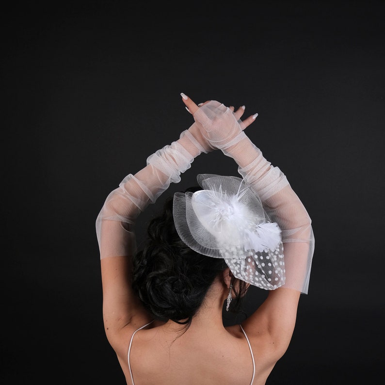 May include: A bride with her hair up in a bun, wearing a white veil with polka dots and a white feather. She is wearing white gloves and has her arms raised above her head.