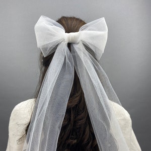 Op de afbeelding: Een witte tule strik met een lange sluier eraan. De strik is in een grote, losse knoop geknoopt en de sluier drapeert over de achterkant van het hoofd.