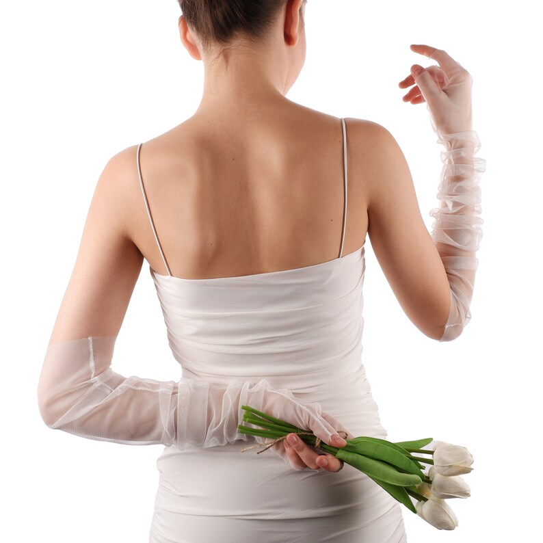 May include: A woman in a white dress and long white gloves holds a bouquet of white tulips behind her back.