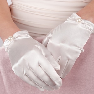 May include: A pair of white satin gloves. Each glove has a pearl accent at the wrist. The gloves are smooth and appear to be new. The background is a light pink and white.
