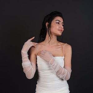May include: A woman wearing a white sleeveless dress with a ruched bodice and long white mesh gloves. She is looking to the right and has her hair pulled back.