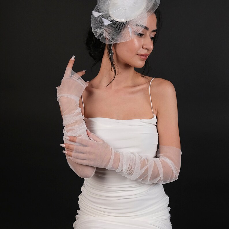 May include: A white tulle dress with a strapless neckline and a white tulle hat with feathers. The model is wearing white tulle gloves.