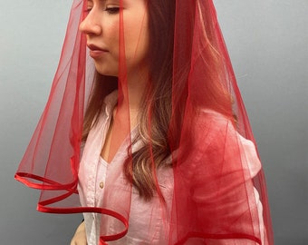 May include: A red tulle veil with a satin trim. The veil is worn by a person with long brown hair and a crown of pearls and flowers.