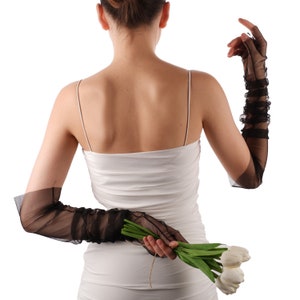 May include: A woman wearing a white sleeveless top and black sheer gloves holds a bouquet of white tulips behind her back.