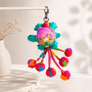 Peut inclure: Un porte-clés en forme de tortue colorée avec une carapace à motifs floraux et des nageoires turquoise. Le porte-clés a un fermoir en métal et comporte des brins pendants avec des pompons de différentes couleurs. La carapace de la tortue est rose vif avec des motifs floraux jaunes et bleus.