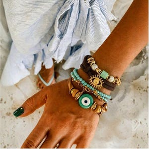 Puede incluir: Una colección de pulseras doradas y turquesas adornan una muñeca. Las pulseras presentan un dije de ojo turco, cuentas y una envoltura de cuero marrón. La mano tiene una manicura verde. El fondo muestra a una persona con una prenda a rayas azules y blancas.
