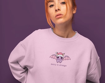 May include: Pale pink sweatshirt with a cartoon bat wearing a Santa hat, and the words "merry & strange" underneath. The bat has purple wings and a smiling face. The sweatshirt is worn by a person with red hair.