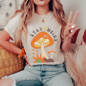 May include: Beige t-shirt with a retro mushroom graphic and the text "Stay Weird." The design features an orange mushroom with yellow and white details, along with smaller mushrooms and flowers. The shirt is paired with light blue jeans.