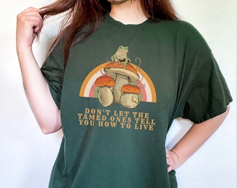 May include: Dark green t-shirt featuring a whimsical design of mushrooms, a frog, and a rainbow. The text "DON'T LET THE TAMED ONES TELL YOU HOW TO LIVE" is printed below the graphic. The shirt is a relaxed fit.